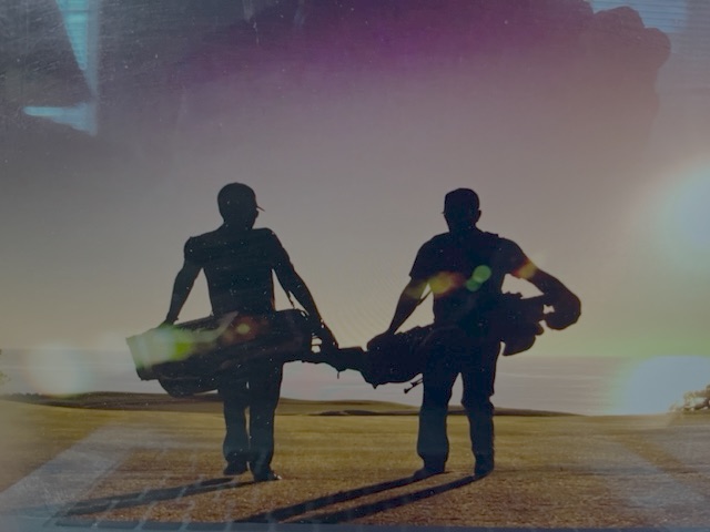Two caddies carrying golf bags walking a fairway at sunset &mdash; &ldquo;The Caddie Chronicles&rdquo;